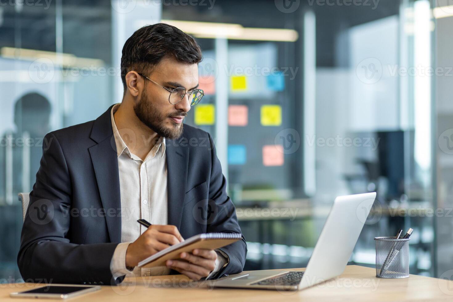 A focused businessman, clad in a suit, meticulously takes notes while seated at a desk, utilizing a laptop in a modern office setting. photo