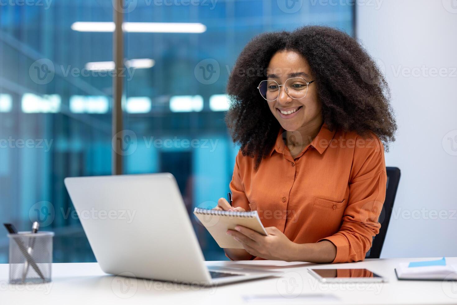 A smiling young woman takes notes during a call in an office setting. She's using her laptop and wearing glasses. photo