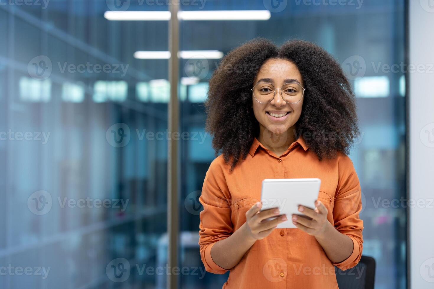 A smiling African American woman with glasses holds a tablet in an office setting, suggesting digital engagement and modern work. photo