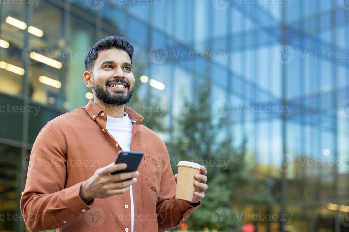 A smiling Indian man holds a coffee cup and smartphone in front of a modern glass building on a sunny day. photo