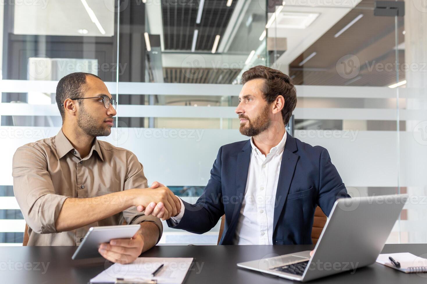 Two businessmen shaking hands in a modern office setting, likely after a successful deal or agreement. Focus on collaboration and teamwork. photo