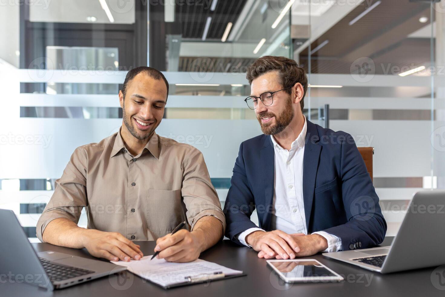 Two businessmen are sitting at a table, one is signing a document, while the other watches. They seem happy. photo
