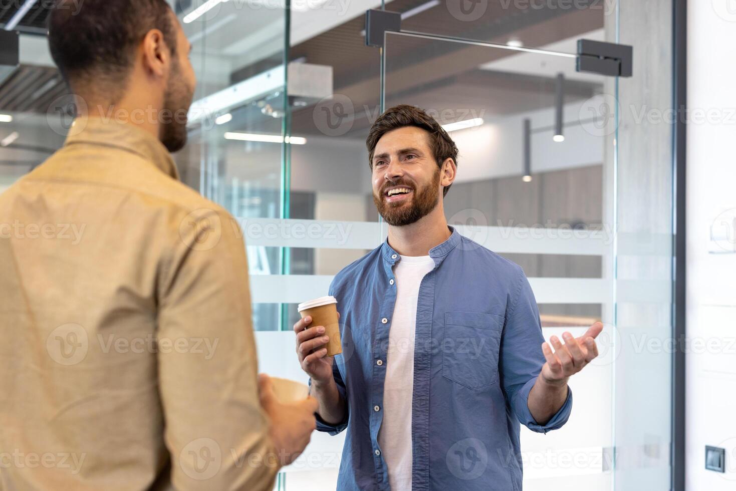 Two men are chatting in an office, one holding a coffee cup and gesturing while the other listens. photo