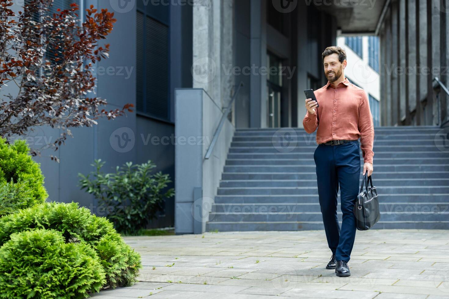 Successful businessman walking through the city with phone in hands and business bag. Man in shirt typing text message and browsing social networks in full growth. photo