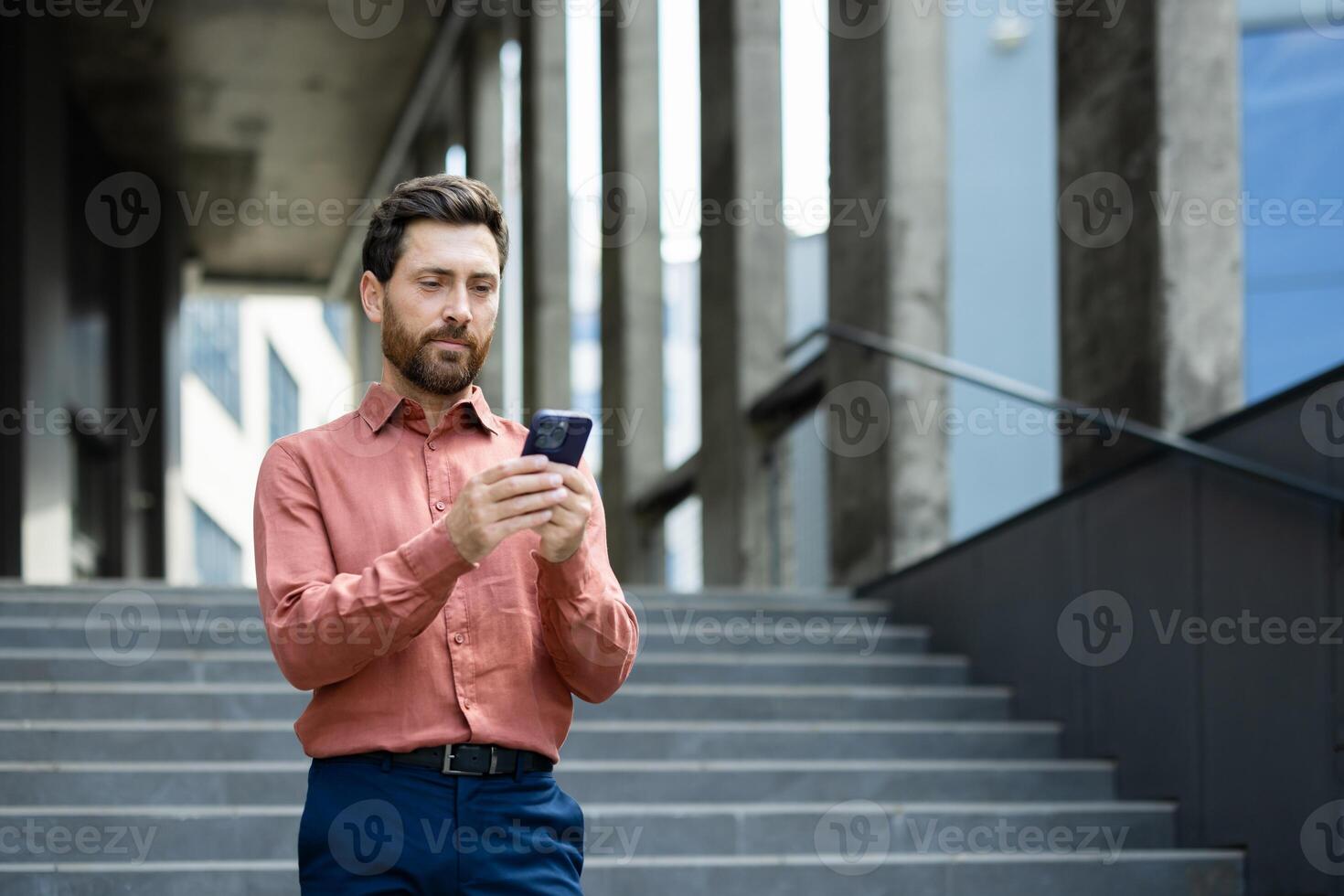 Serious thinking businessman with phone in hands reads online messages and browses internet pages. Man uses smartphone outside office building, walking through city. photo