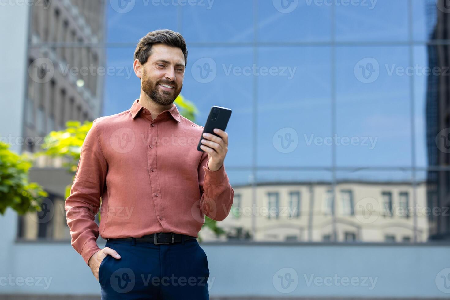 A smiling businessman in a casual outfit looks at his phone in front of a modern building. The man appears to be outside. photo