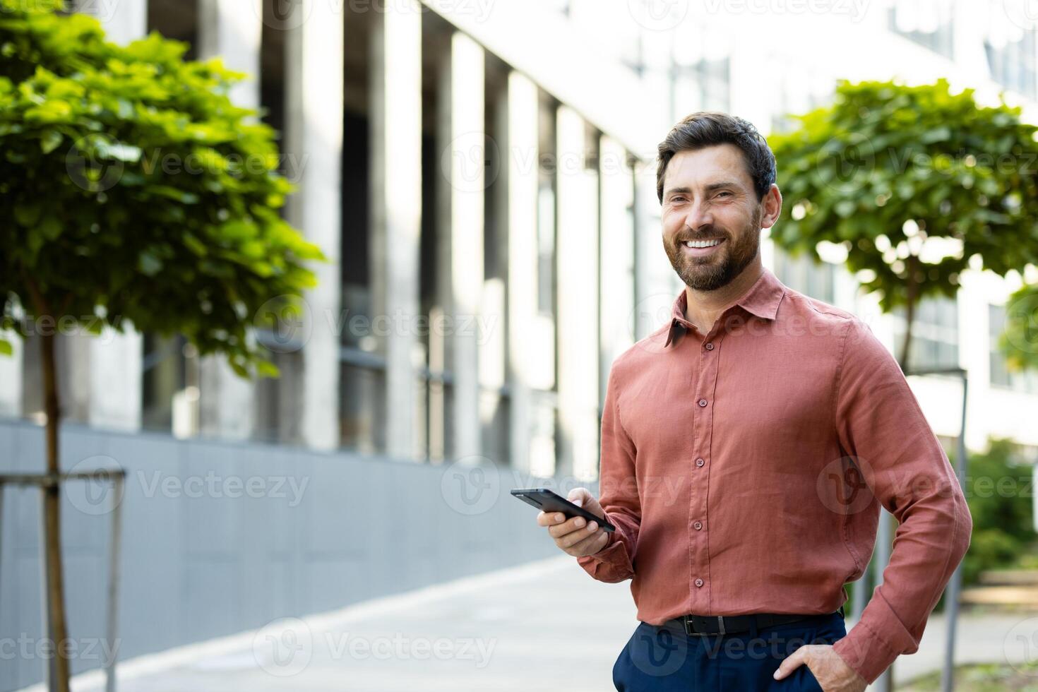 Portrait of a successful and joyful businessman. A man with a phone in his hands is smiling and looking at the camera while walking outside an office building. photo