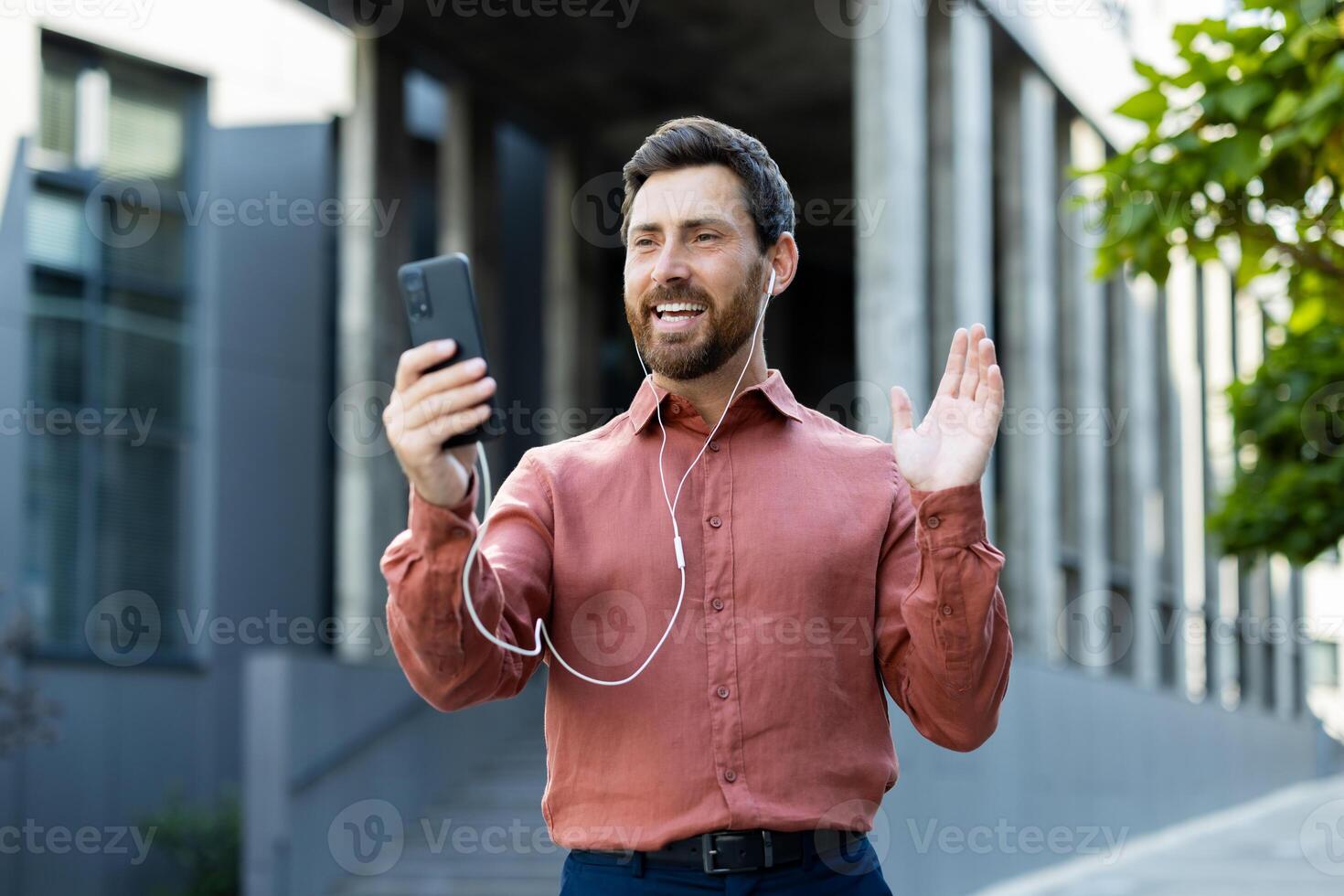 Smiling happy man using phone and wired headphones for call. Businessman talking to colleagues and partners remotely while walking outside office building, city. photo
