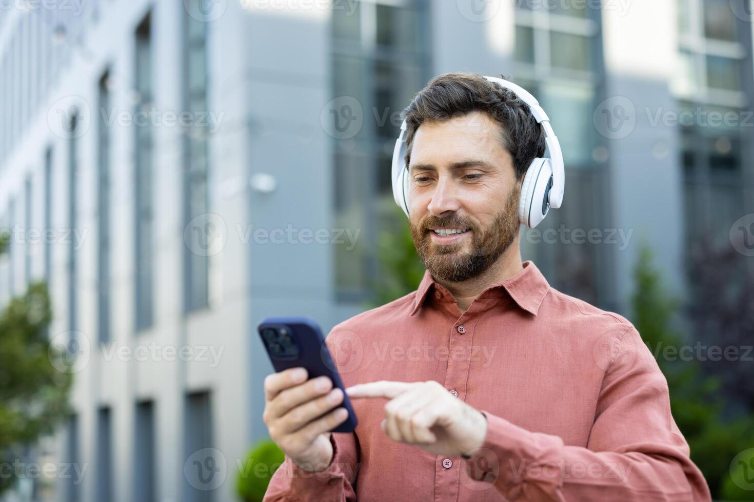 A man with headphones uses his phone outside on a city street with modern buildings, enjoying music or connecting with others photo