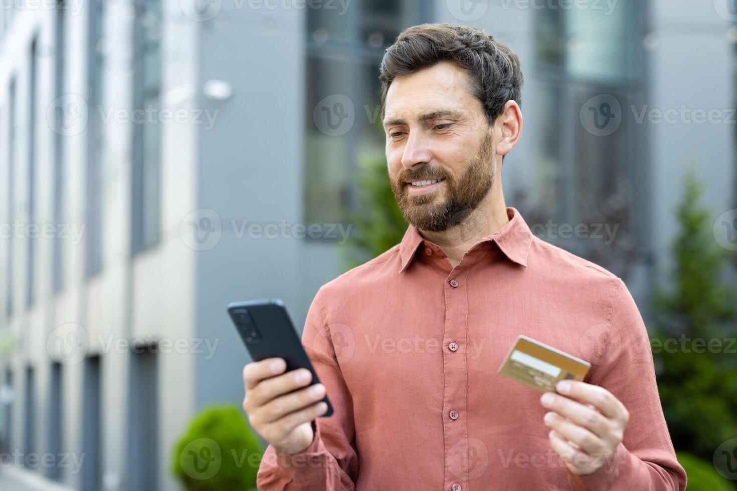 A man smiling while holding a credit card and phone, likely making an online purchase outdoors. photo