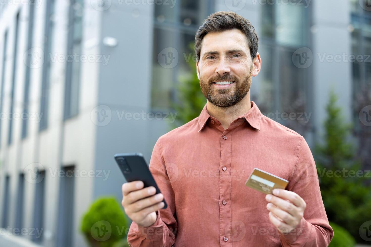 Portrait of happy joyful male shopper with phone in hands and bank credit card. Businessman smiling and looking at camera, walking happily outside office building in city photo