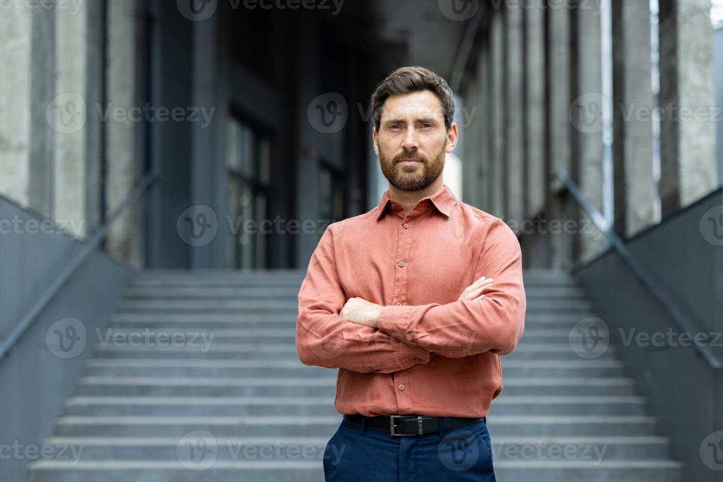 Portrait of confident mature businessman with crossed arms, standing outdoors against modern building. He has a beard and is wearing a terracotta shirt. photo
