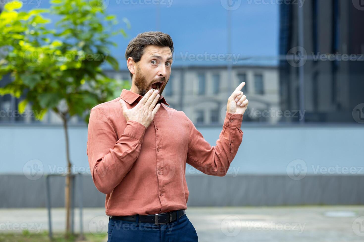 A surprised man in a shirt points upwards with an open mouth and a hand to his mouth, standing outdoors with a modern building behind. photo