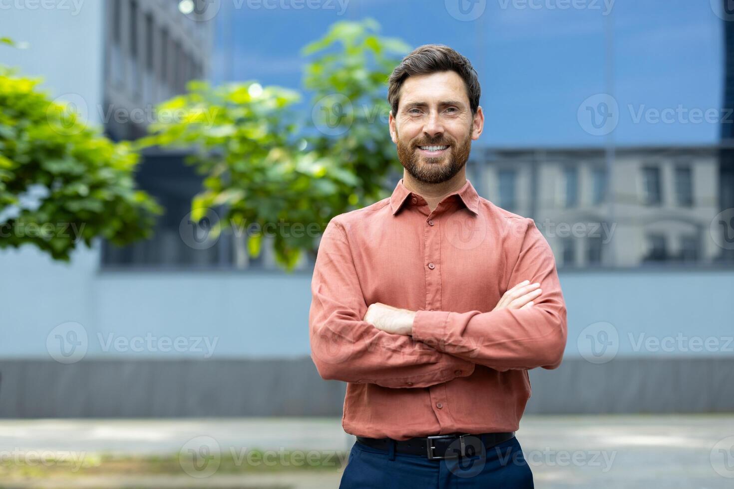 Portrait of successful businessman in shirt, man smiling and looking at camera with crossed arms outside office building. Satisfied with results of achievement, company employee. photo