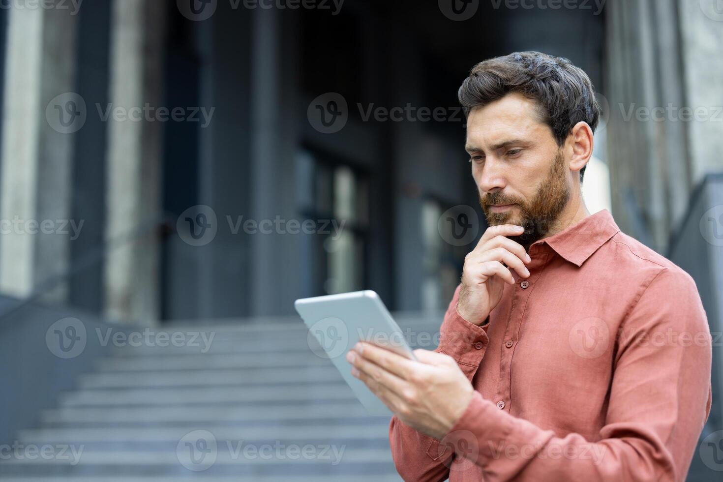 A contemplative man with a beard reviews data on a tablet device in front of a modern, gray building, looking thoughtful and engaged. photo