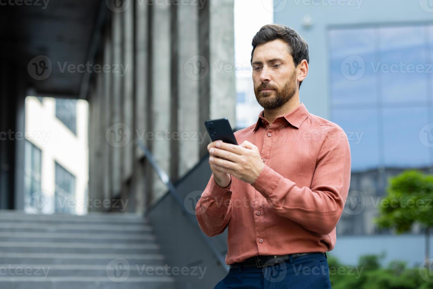 A handsome man in a shirt checks his smartphone outside of a modern building, likely communicating with colleagues or clients photo