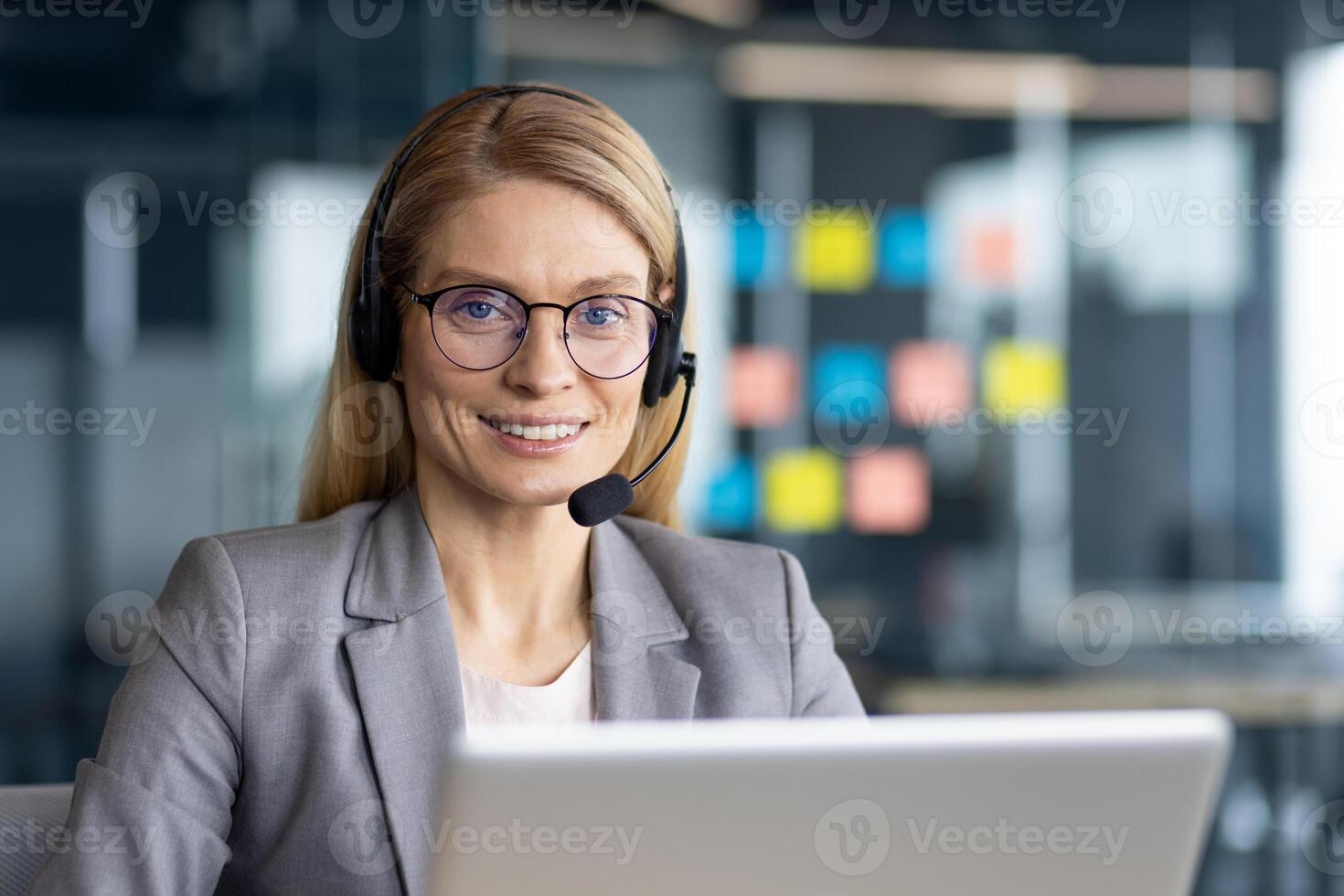 A friendly blonde woman with glasses wears a headset, smiling at the camera while working on a laptop in an office setting. photo