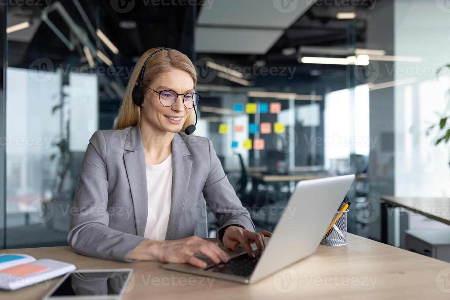 A smiling businesswoman wearing glasses and a headset uses a laptop in a modern office, possibly providing customer service or working remotely. photo