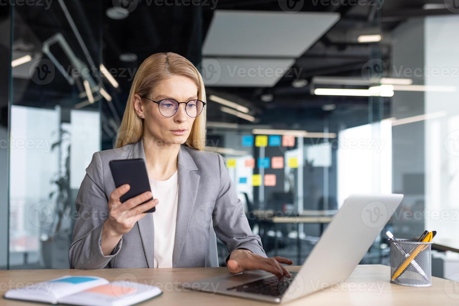 A businesswoman in a modern office setting uses her phone and laptop simultaneously, appearing focused on her work. photo
