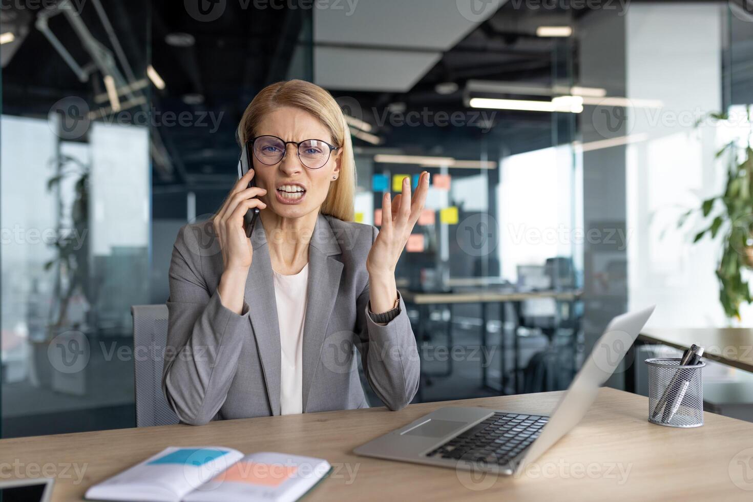 An angry businesswoman in a modern office is on a phone call, looking frustrated and stressed. She is gesturing with her hand and wearing glasses. photo