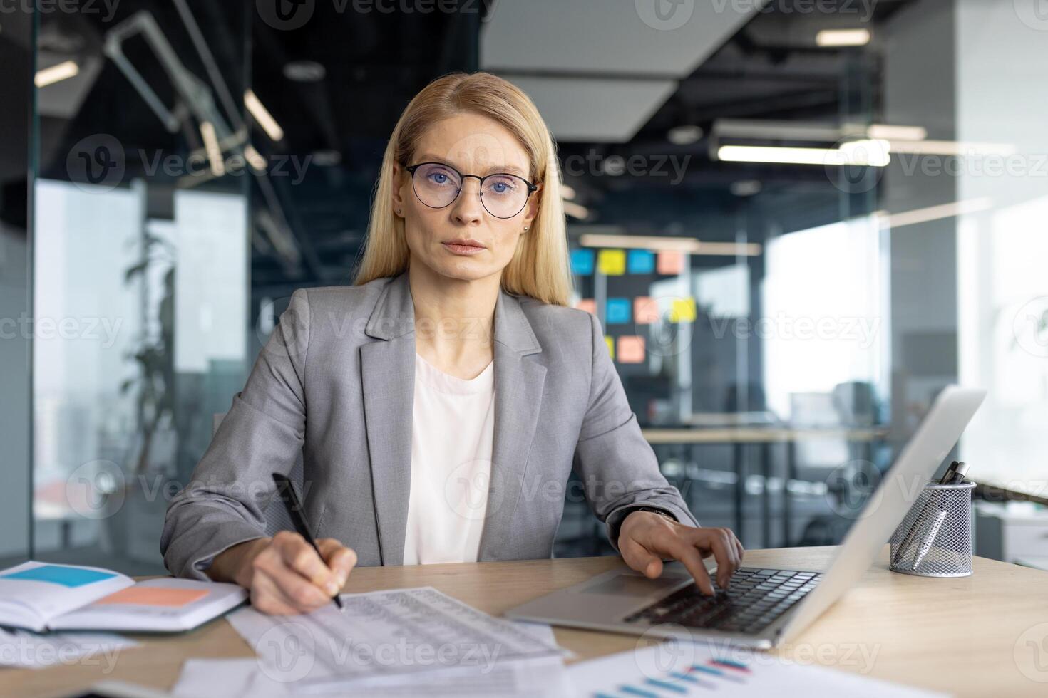 A professional businesswoman in a modern office setting, working on a laptop and taking notes, with a serious expression. photo