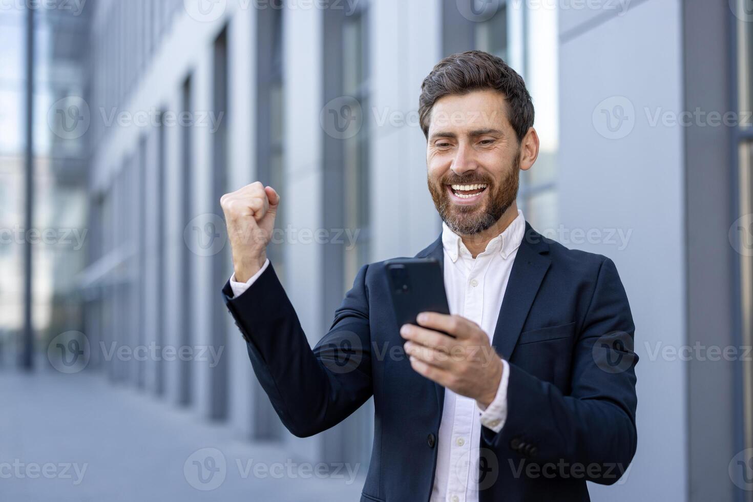 A successful businessman celebrates a win, looking at his phone, with a raised fist and a joyful expression near a modern building. photo