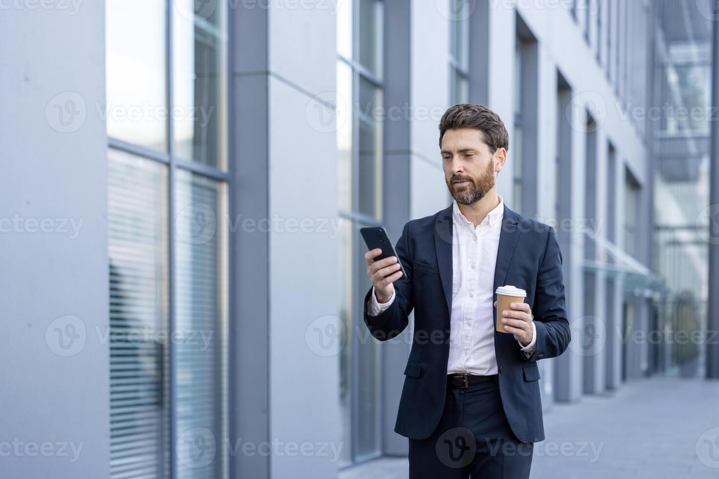 A man in a suit reviews his phone while walking outdoors, holding a coffee cup. The background shows a modern office building. photo
