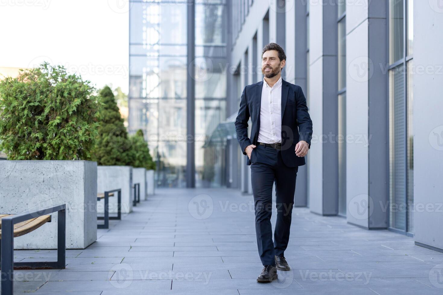 A well-dressed businessman walks confidently in a modern urban setting, exuding professional ambition. He is wearing a dark suit. photo