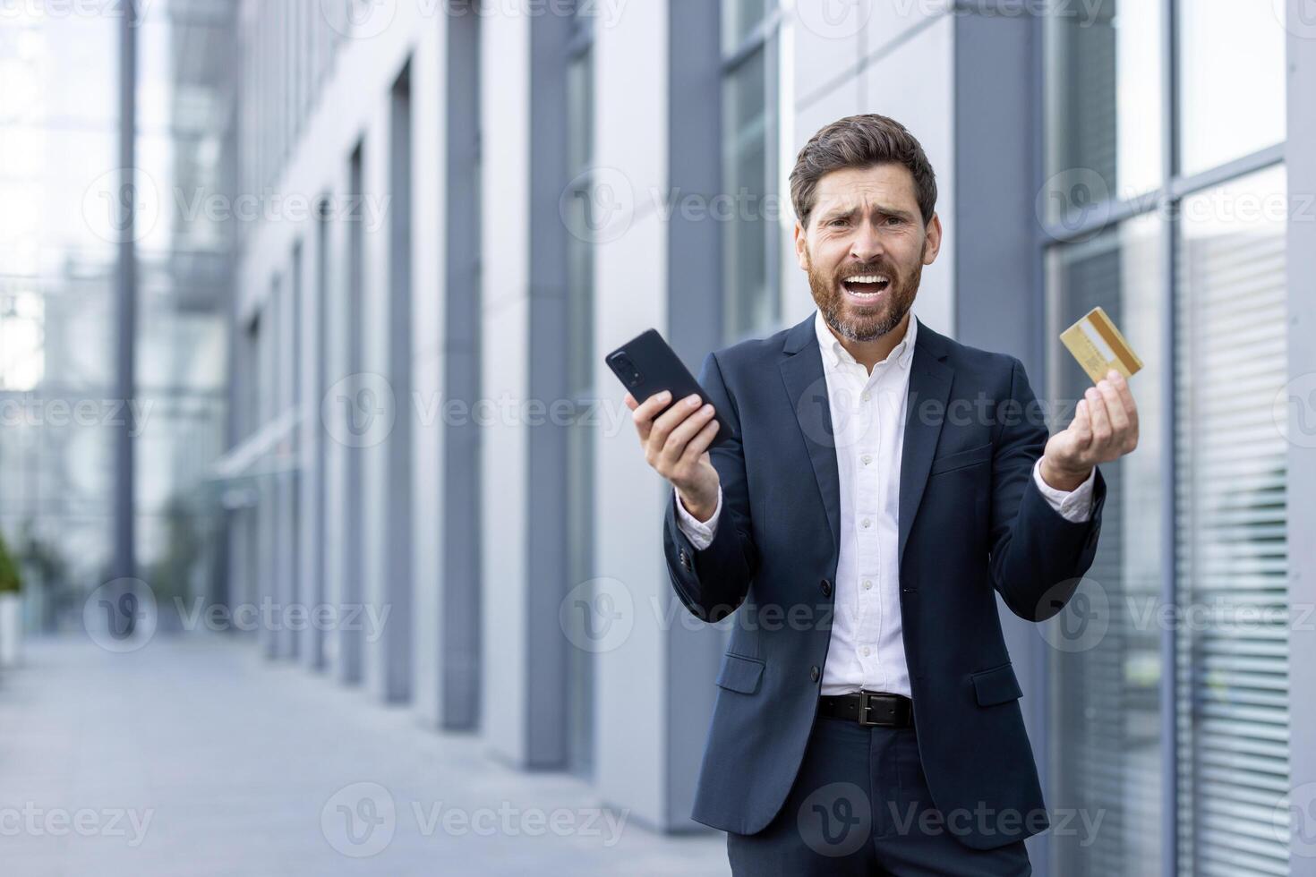 A frustrated businessman, holding a smartphone and a credit card outside an office building, appears to be in a stressful situation. photo