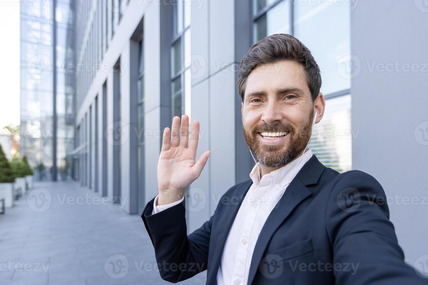 A smiling man waves at the camera while wearing a suit and earpieces, outside a modern building. It's a self-portrait. photo