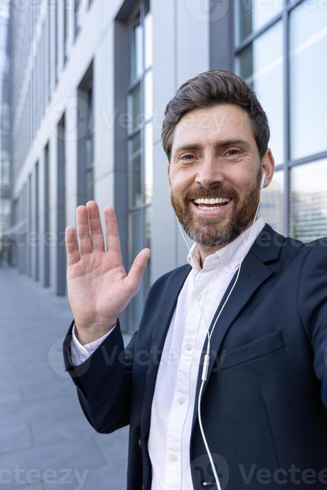 A smiling businessman in a suit waves towards the camera while wearing earbuds outdoors. The background shows an office building. photo