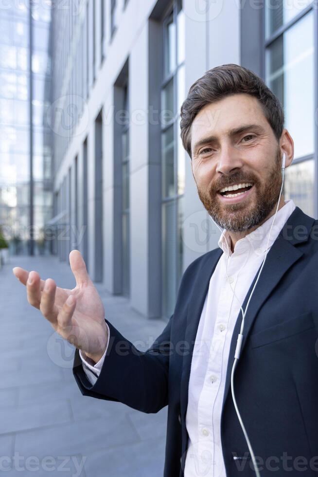 A smiling businessman is on a call in front of a modern building, wearing a suit and using earphones, presenting with his hand photo