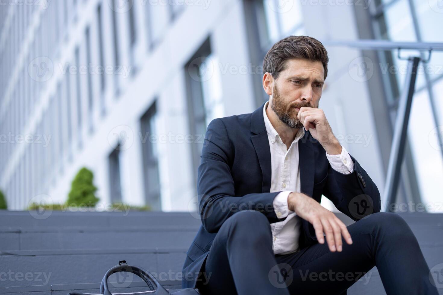 A pensive businessman in a suit sits outside, deep in thought. His expression suggests stress or worry in a corporate environment. A briefcase sits nearby. photo