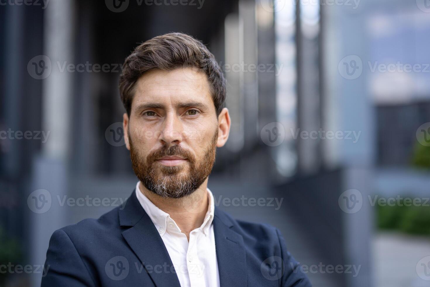 A handsome, bearded man in a suit poses confidently, looking directly at the camera with a composed expression. The urban backdrop hints at a professional setting. photo