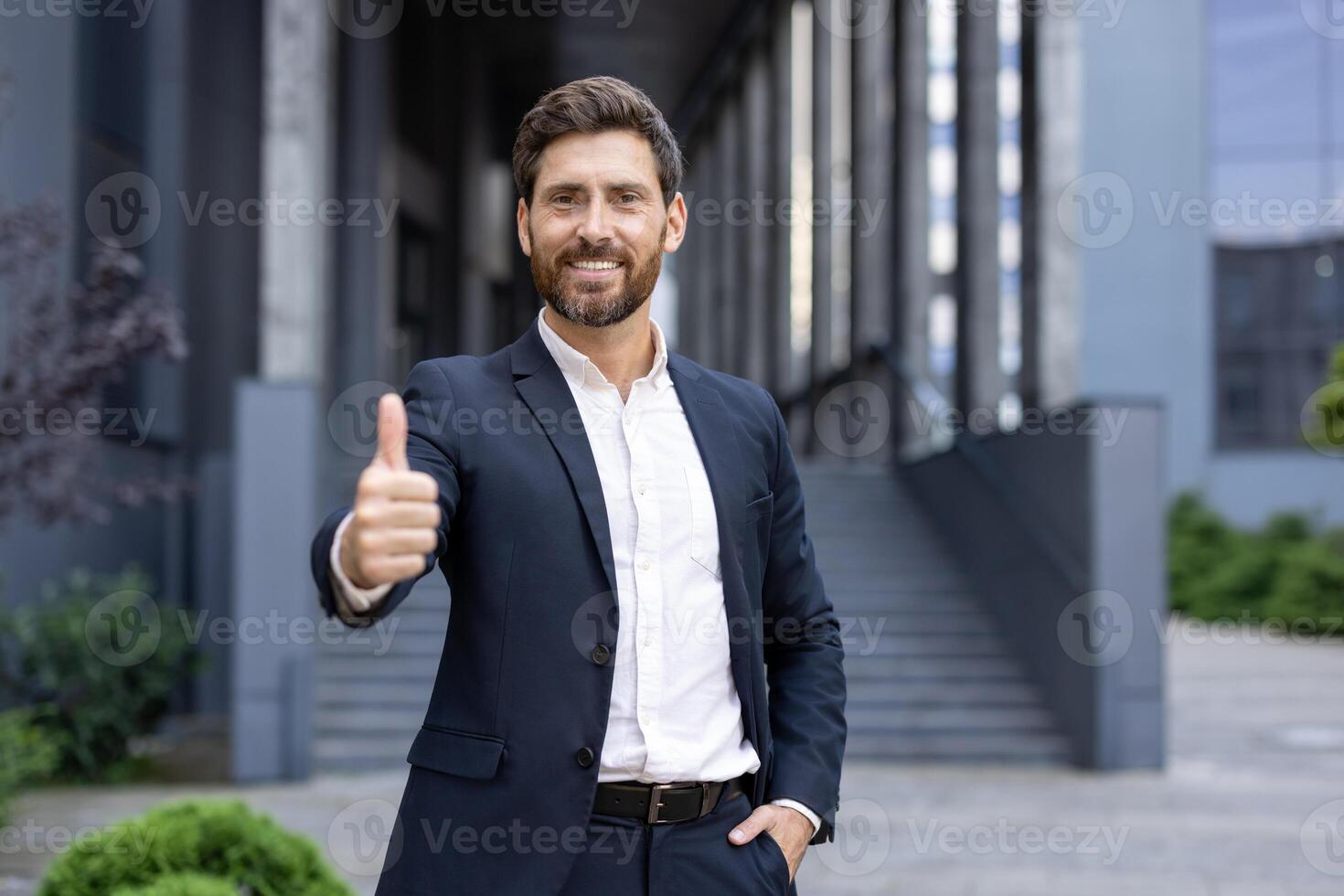 A smiling businessman in a suit, giving a thumbs-up gesture while standing outside of a modern building. photo