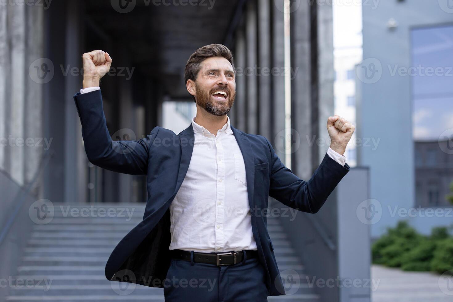 A successful businessman celebrates with his arms raised in front of a modern building, showcasing achievement and triumph. photo