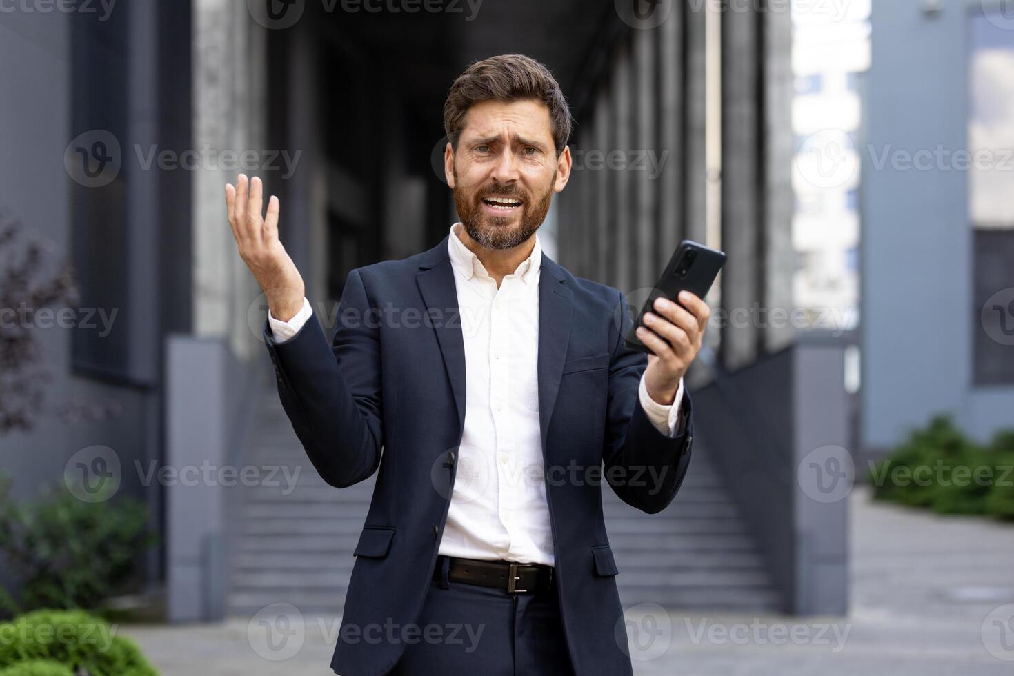 A frustrated businessman in a suit gestures while holding a phone, likely experiencing a work issue outdoors. The city building is the backdrop. photo