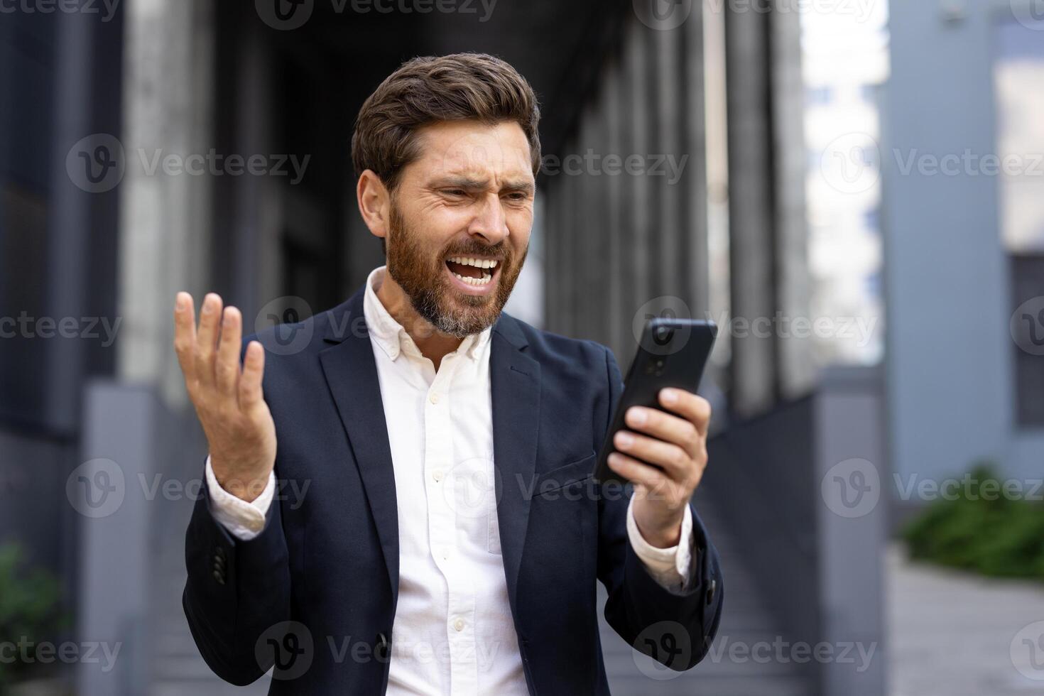 An angry businessman is shown looking at his phone, expressing frustration. He is standing outside a modern office building. photo