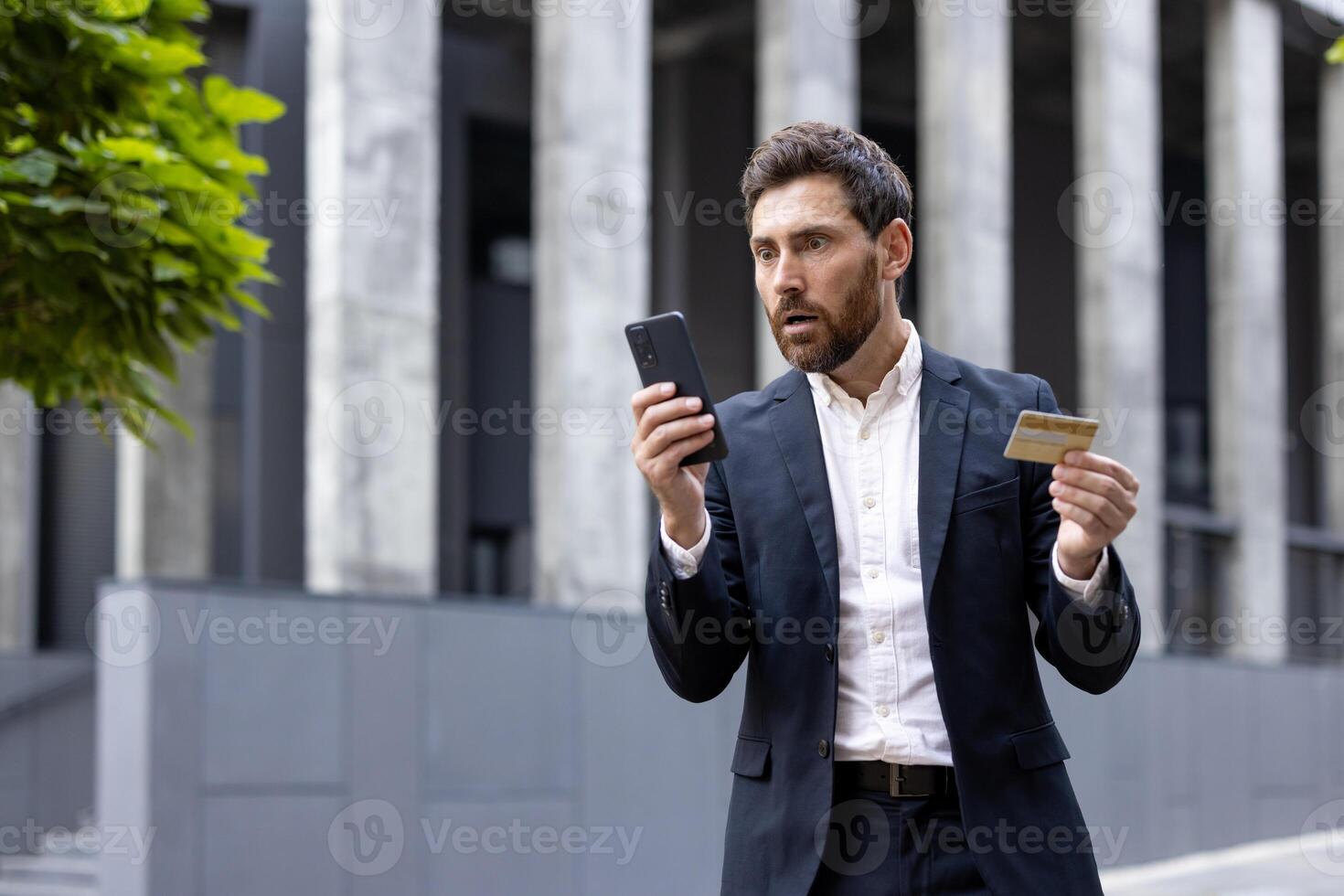 A businessman looks shocked while holding a credit card and his phone, possibly due to a financial issue. photo
