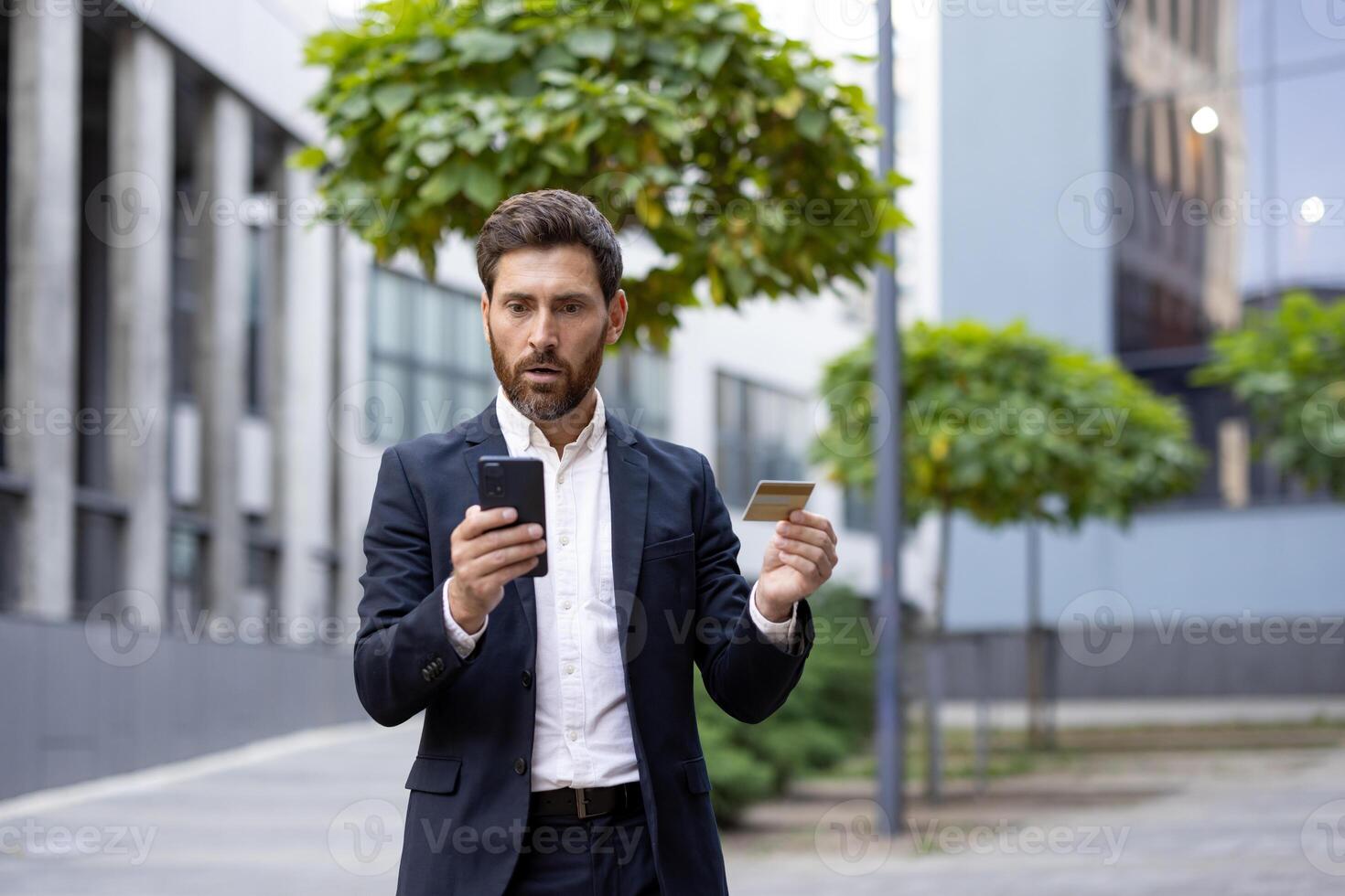 A businessman, looking shocked, holds a credit card and smartphone outdoors, likely dealing with a financial issue. photo