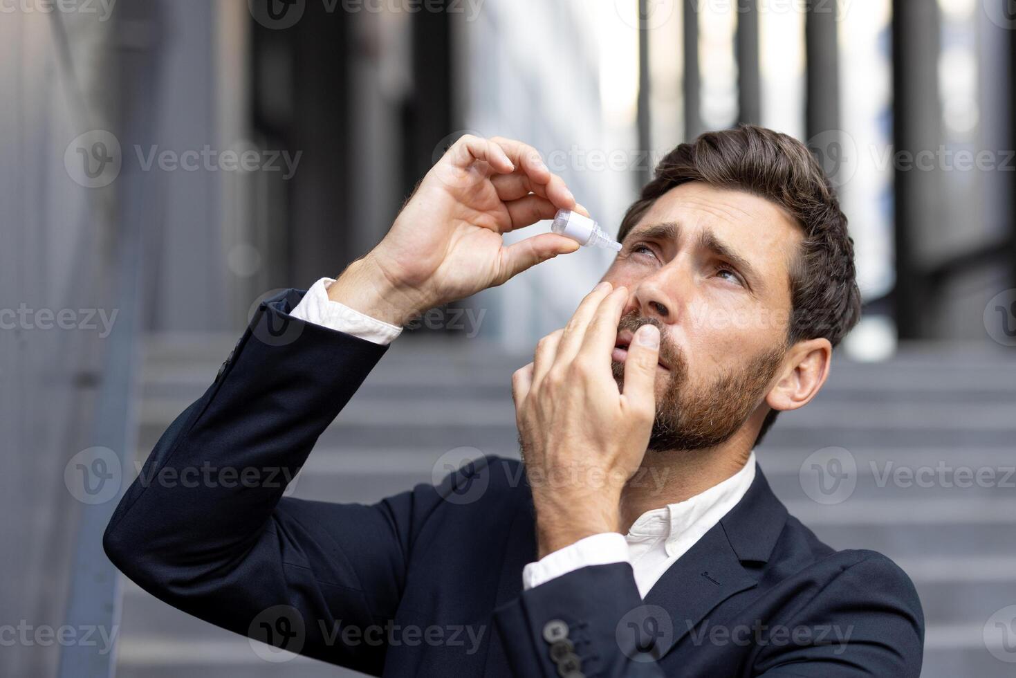 A businessman applies eye drops to soothe irritated eyes. He stands outdoors near office buildings, showing the challenges of modern life. photo