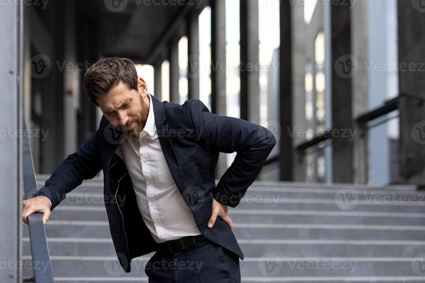 A businessman grimaces in pain as he clutches his back while standing on stairs, highlighting a potential work injury or health concern. photo