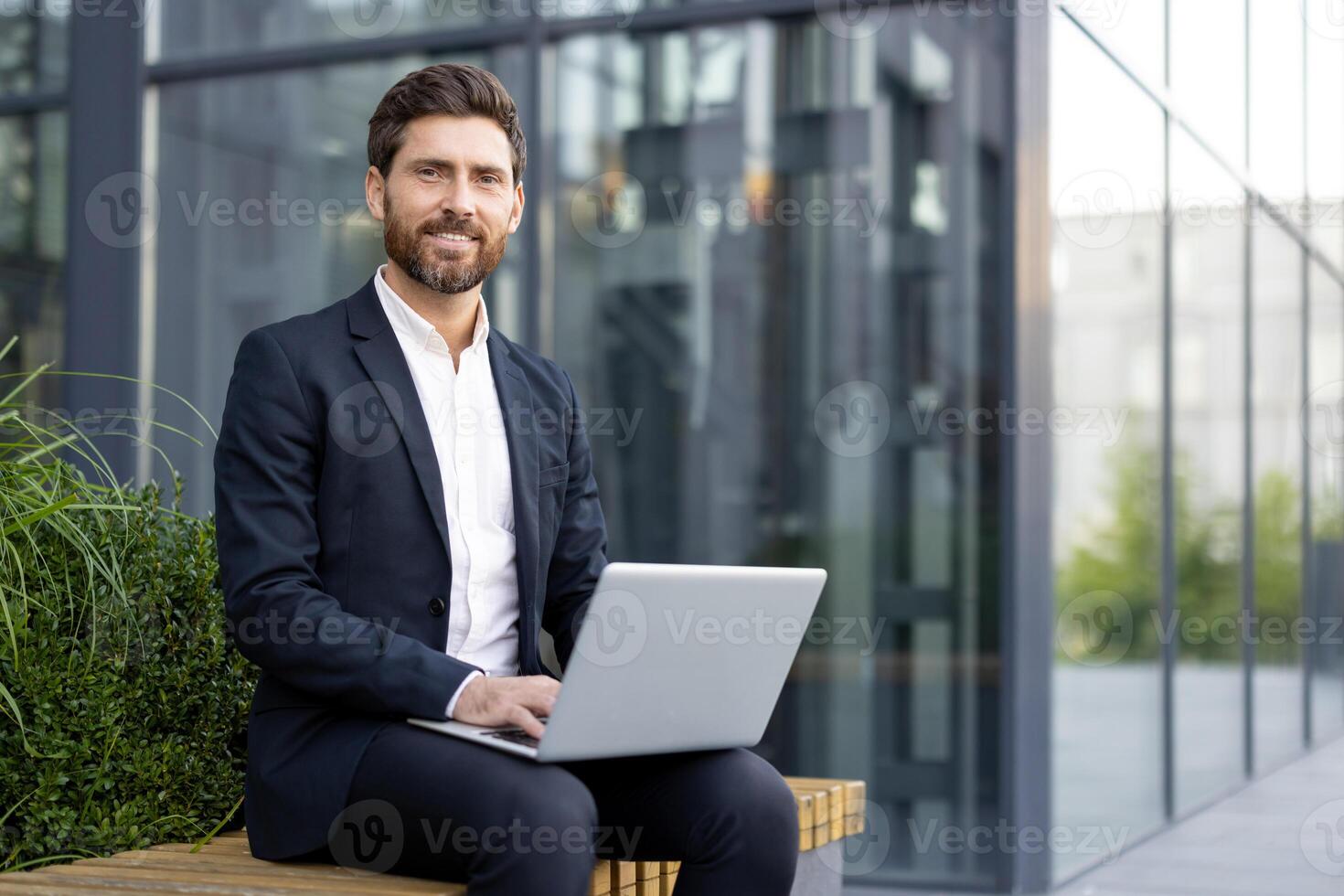 A smiling businessman sits outside, working on a laptop. He wears a suit and is positioned in front of a modern building, suggesting a professional setting. photo