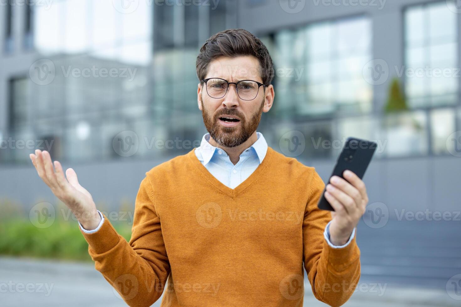 A puzzled businessman holds his phone with an open palm, showing confusion. He stands outside a modern building wearing glasses. photo