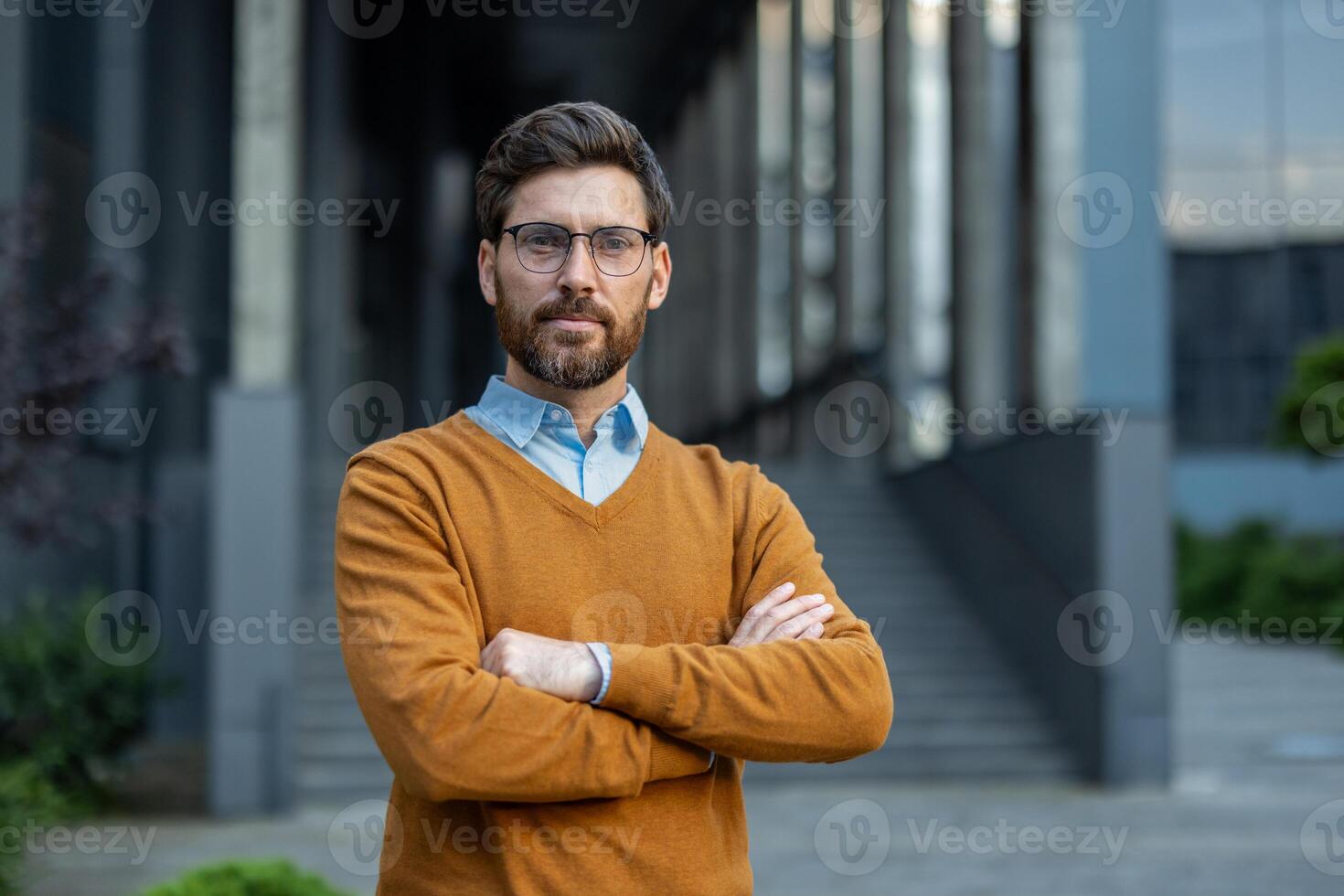 A confident man with glasses stands with arms crossed in front of a modern building, outdoors. photo