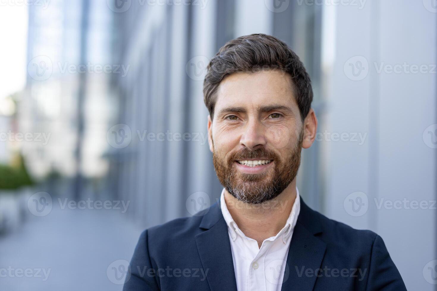 A smiling businessman poses outside of an office building. He is wearing a suit and looking at the camera with a friendly expression. photo