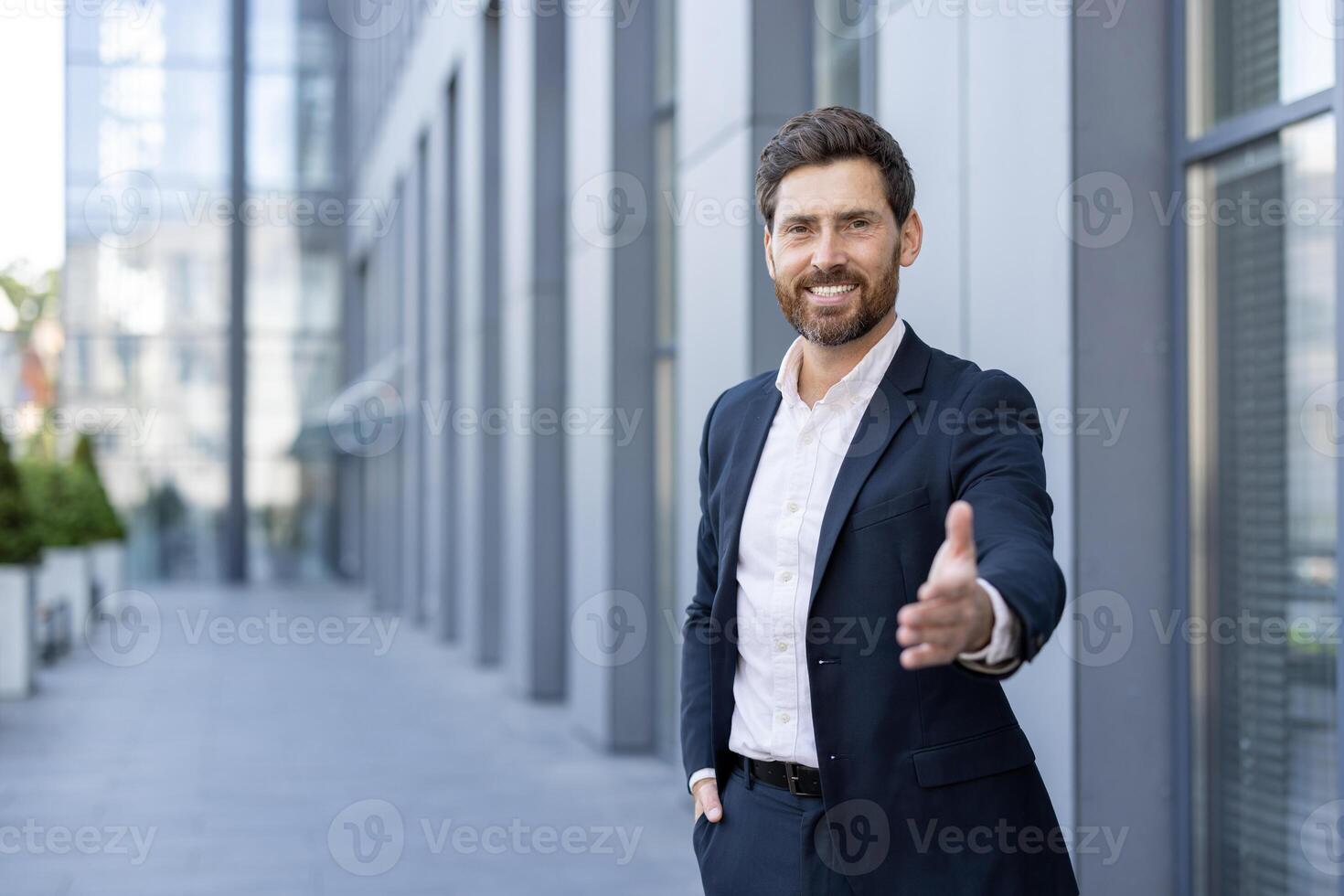 A businessman in a suit offers a handshake, smiling in front of a modern office building. photo