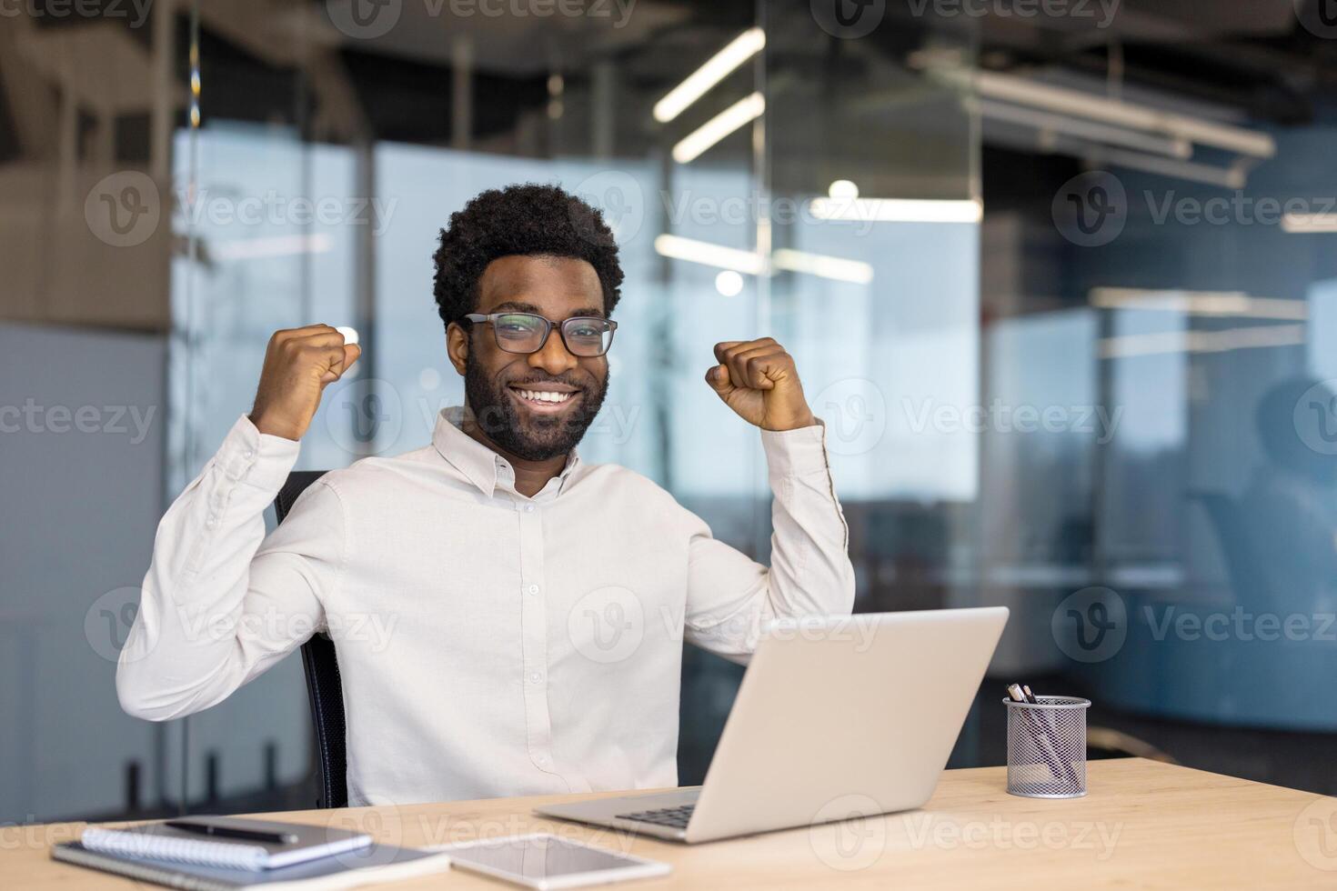 A smiling, happy, man celebrates with raised fists while working on a laptop in an office environment. photo