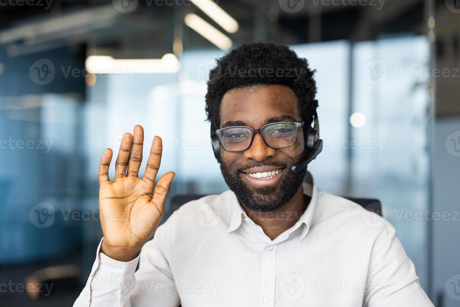 A cheerful man with glasses and a headset waves, expressing a friendly welcome or greeting. He smiles in an office setting. photo