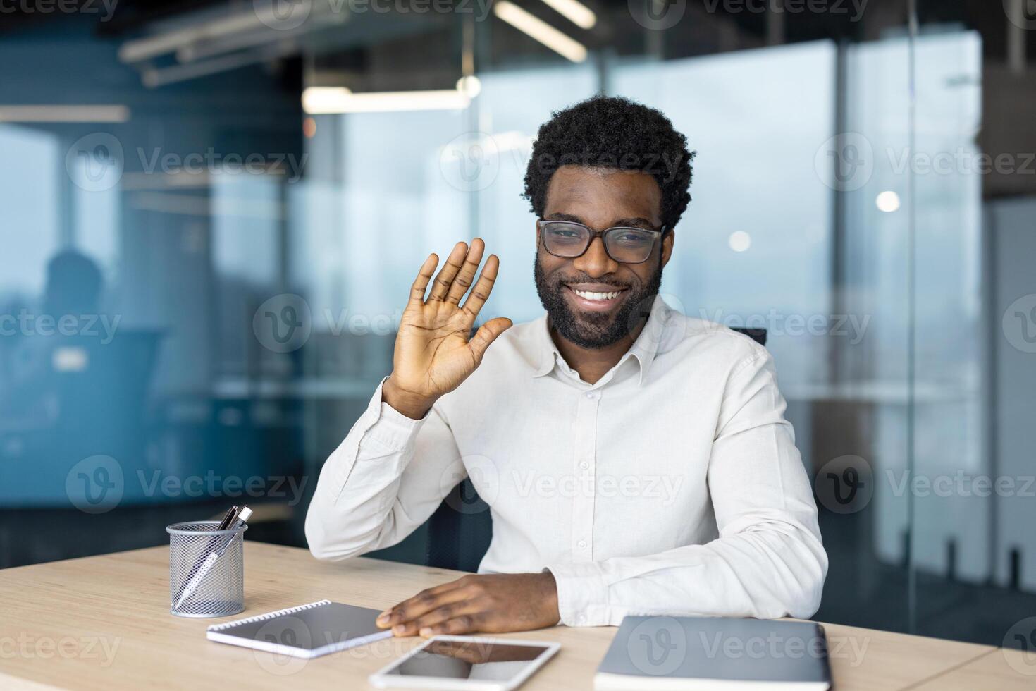 An African-American man in glasses waves and smiles while sitting at a desk in an office. He appears happy and approachable. photo